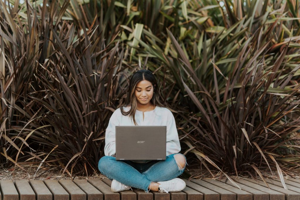 A woman in a white shirt and blue jeans sits on a boardwalk next to tall green and brown leafy plants. She's on a silver Acer computer, possibly looking up AI wedding planners.