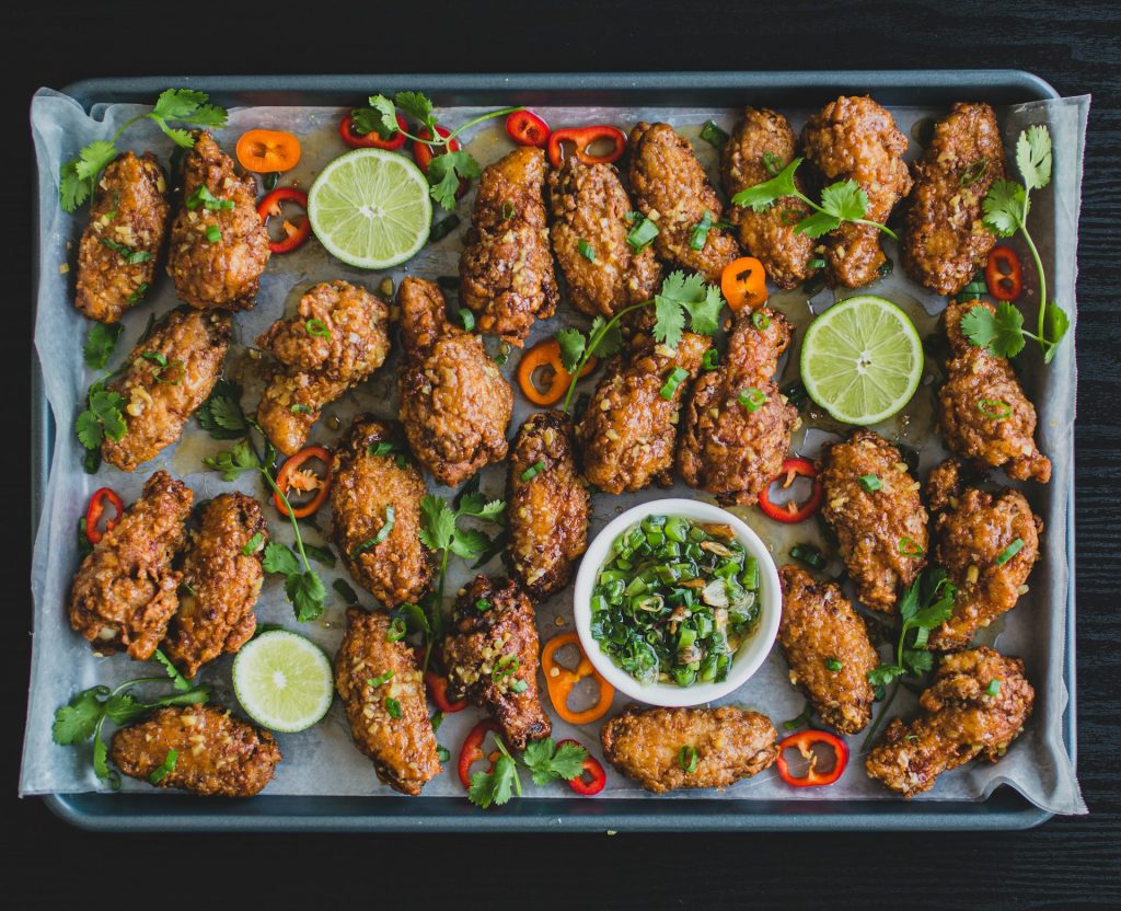 A baking sheet lined with wax paper sits on a dark brown table. It's covered in fried chicken wings, sliced mini bell peppers in orange and red, sliced limes, a chive-based dip and garnished herbs. The wings are one of the air fryer hacks people like to make.