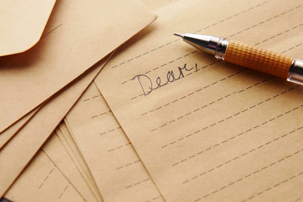 A silver and orange pen rests on a nearly blank sheet of paper. The word "Dear," is written in cursive. Other blank sheets of the same paper lie under the start of the letter.