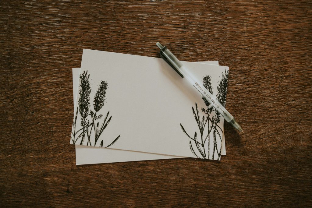 A blank greeting card with black tall flowers sits on a white envelope on a brown wooden surface. A clear and silver pen sits on the card, waiting for someone to learn how to write an apology letter.