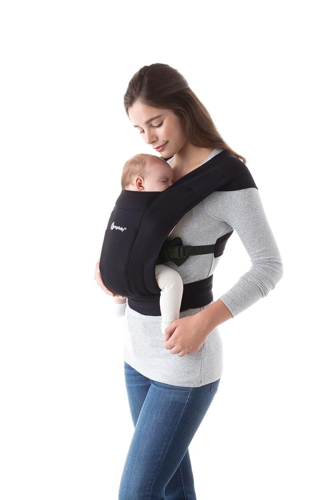 A woman in a gray shirt and jeans wears a newborn carrier wrap with a baby in it.