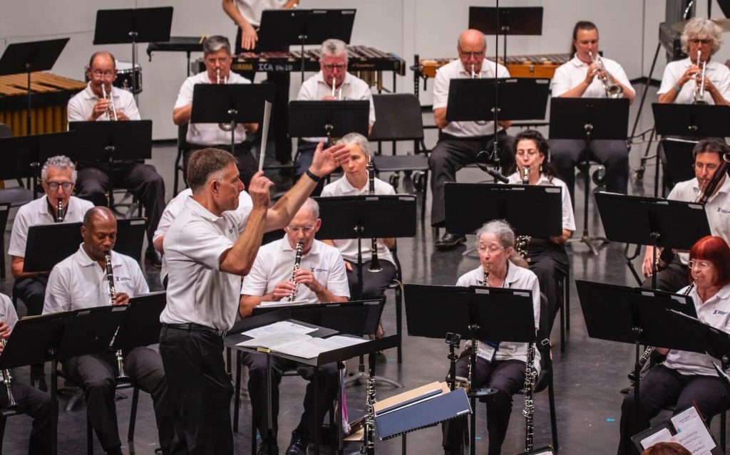 A man in a white polo shirt and black pants directs an orchestra of people in the same uniform. They're rehearsing for a show.