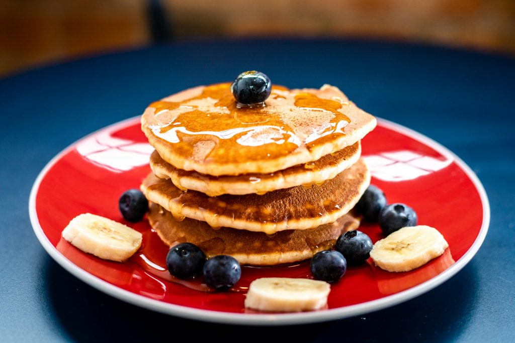 A red plate sits on a blue table, covered in four pancakes, blueberries and banana slices covered in maple syrup.