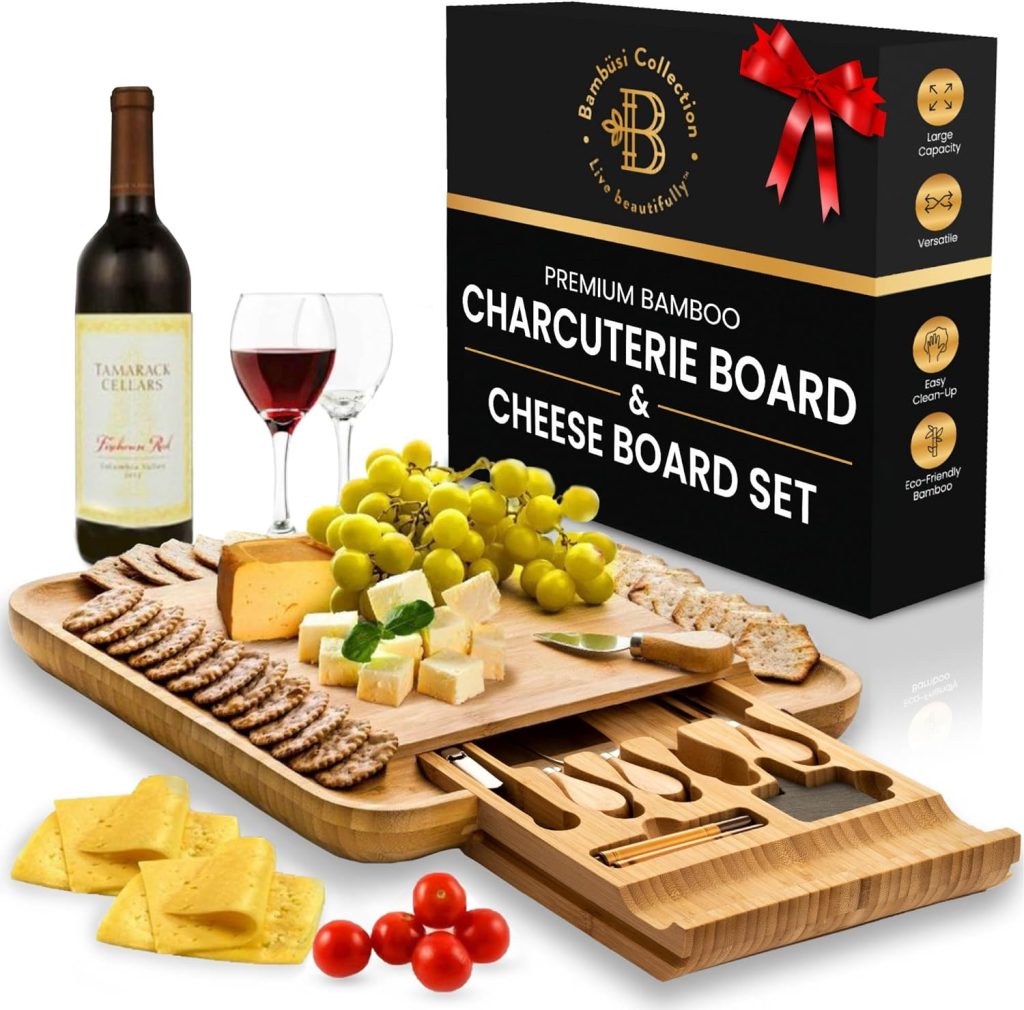 A fancy charcuterie board with multiple levels and a hidden cheese knife drawer is ready for dining, covered in crackers, fruit and cheese. A bottle of red wine is in the background. One wine glass is full and the other isn't. The box that the charcuterie board comes in is standing in the background.