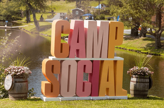 The Camp Social sign with pink and yellow letters sits on a sunny, grassy hill overlooking a lake and the camp.
