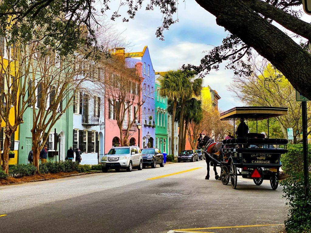 A downtown street in Charleston features multi-colored pastel buildings. A horse-drawn carriage enters one lane in the road while cars are parked on the other side. People walk down the sidewalk against the buildings.