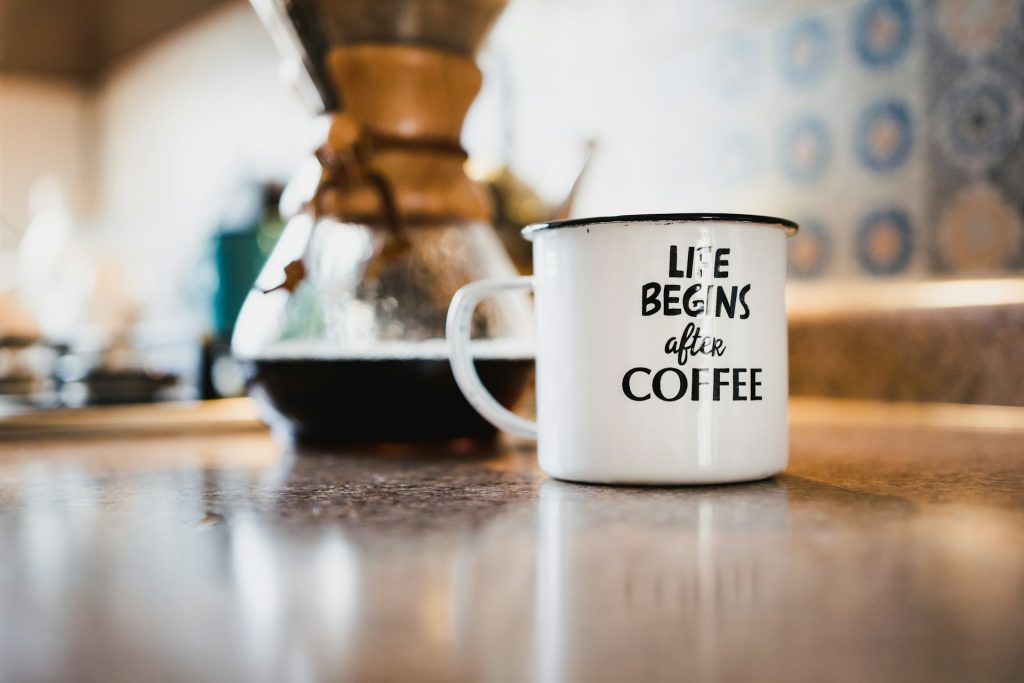 A white mug that says Life Begins After Coffee sits on a brown kitchen counter in front of a press filled with dark coffee.