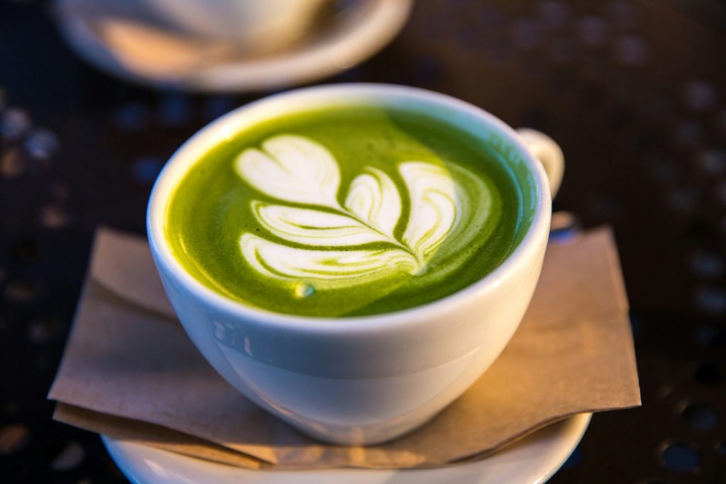 A white mug of matcha tea with a white leaf shape in the top sits on a white plate and a brown napkin. Another sits behind it, likely for people who compared coffee vs. matcha and chose to drink matcha.