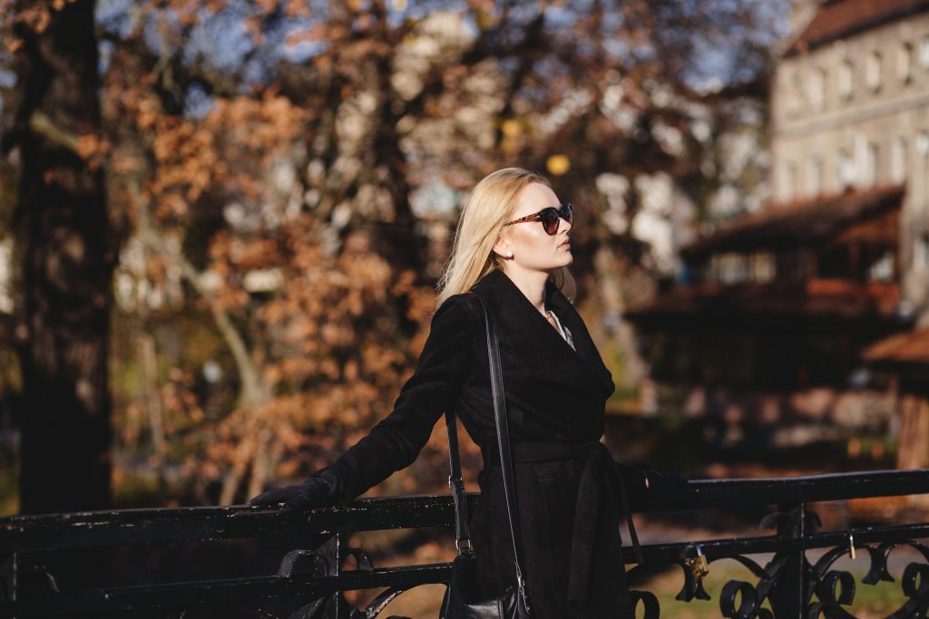 A blonde woman wearing dark sunglasses, black trench coat tied at her waist, a black purse and black gloves stands with her hands on the black iron railing of a bridge. Autumn trees are blurred behind her in addition to a few buildings on the other side of the river she stands over.
