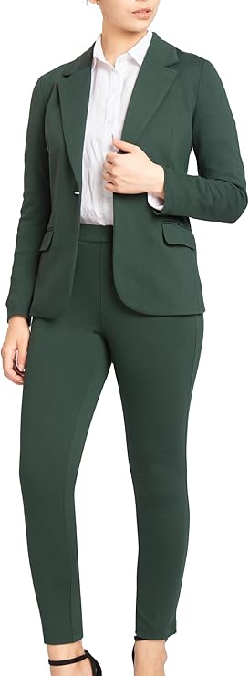A woman stands in a green blazer and pant suit set with a white shirt and black heels against a white background.