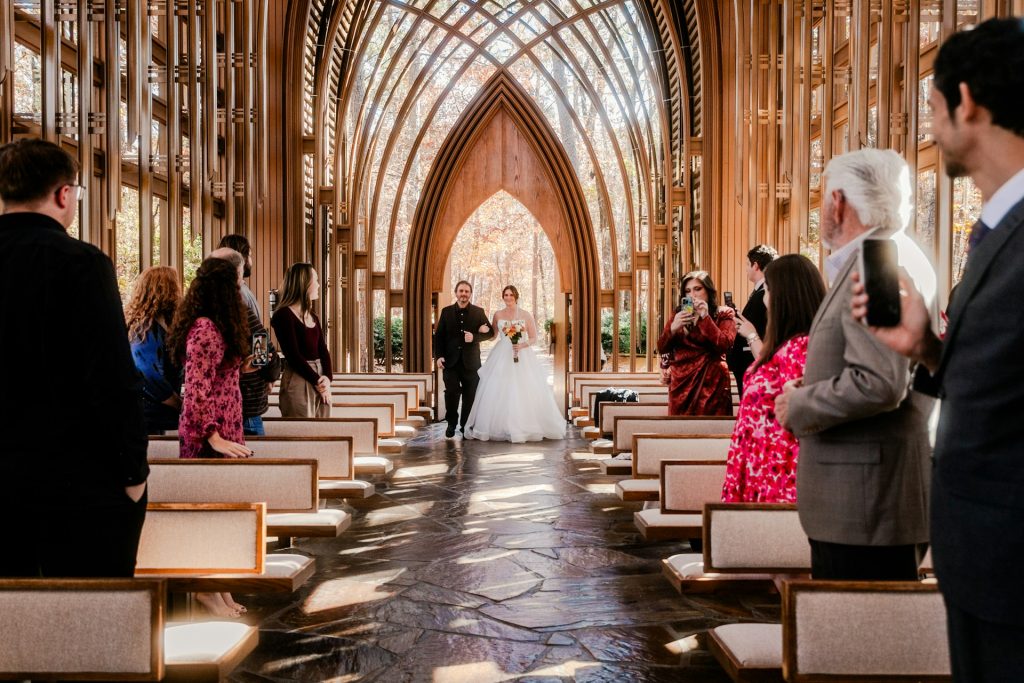 A newly married husband and wife walk back down the aisle of their wedding ceremony while guests in fall wedding guest dresses and other attire take pictures of them. They're in a chapel with stone floors and windowed walls.
