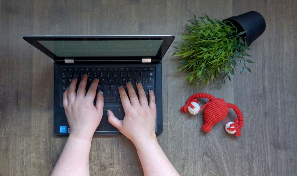 A woman types on a small black laptop on a gray wooden table. A crocheted uterus and fallopian tube plushie in red and white sits next to the computer. A green potted plant in a black pot sits sideways next to it.