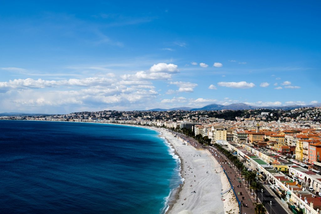A curving coastline of the French Riviera. There's some empty beach space in front of a large city, a background of mountains and a large blue sky with distant white clouds.
