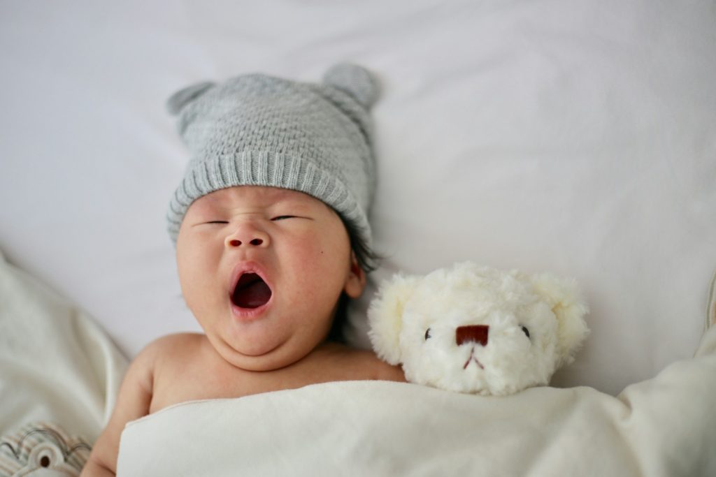 A newborn baby wearing a gray knit hat with two bear ears yawns while tucked under a white blanket. A white teddy bear is slightly smaller than the baby and sits tucked under the same blanket right next to them.