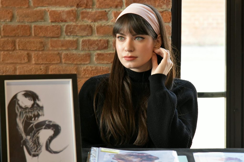 A woman with long brown hair, cat eye eyeliner and a pink fabric headband sits at a table in front of printed pictures. She has curtain bangs that part over her brows. A picture of Venom leans against an exposed brick wall.
