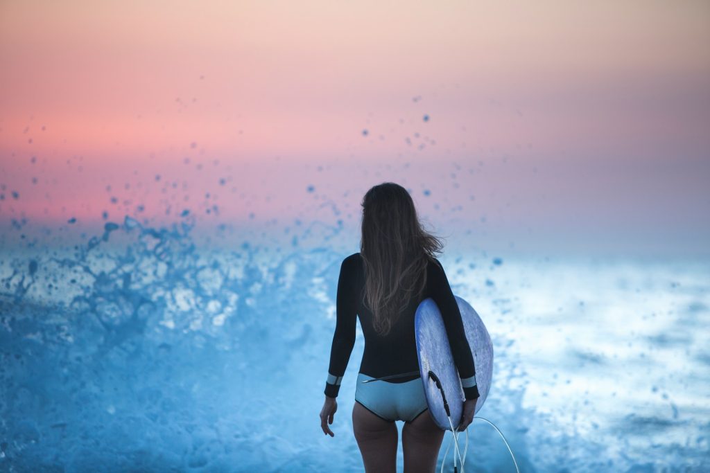 A woman stands in a long-sleeved black swimsuit in front of a crashing wave at sunset. she holds a white surfboard.