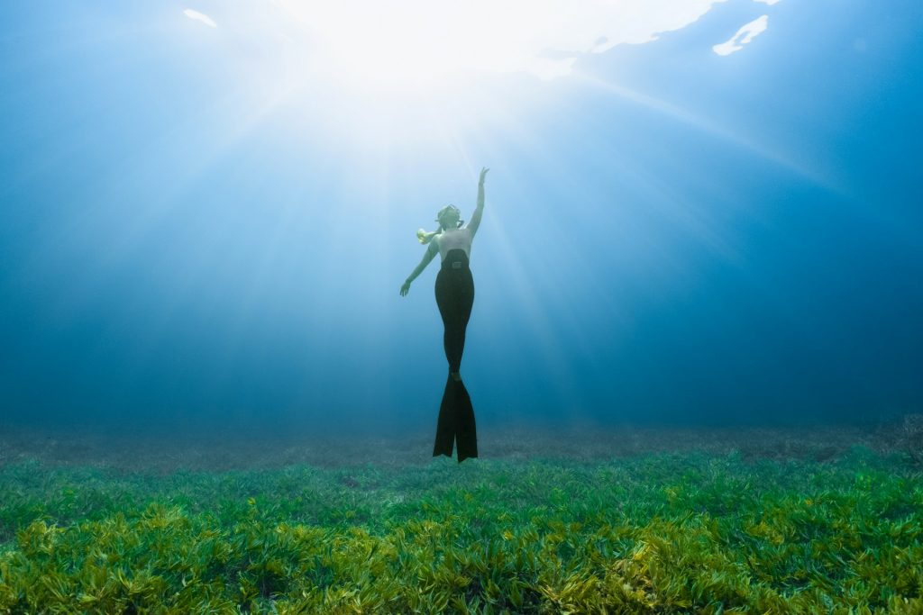 A woman is on the bottom of the ocean where there's lots of kelp. She kicks her feet together to mimic a mermaid while she reaches toward the sun shining above the surface. She wears a snorkeling mask and mouthpiece.