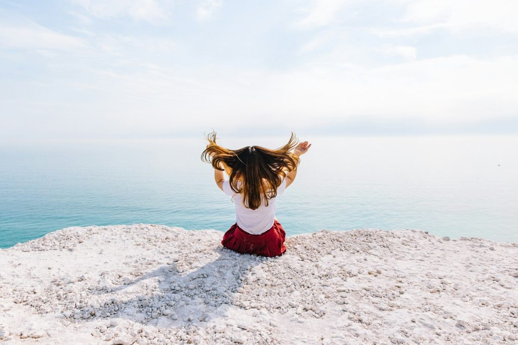 A woman in a white shirt and red skirt sits on a white rocky cliff. She faces the ocean in the distance and flips her hair over her shoulders with both hands.