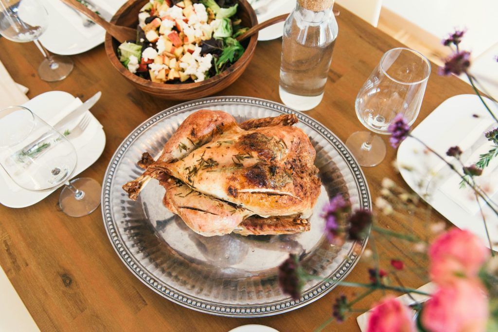 A roasted turkey covered in spices sits on a silver platter on a wooden table surrounded by glasses and bottles of water, a wooden bowl full of salad and white plates with silverware on them. A large floral arrangement of pink and purple flowers sit to the right.