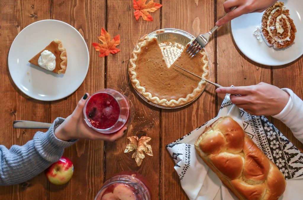 Two friends sit down for Thanksgiving. One is cutting a pumpkin pie with various other desserts on the wooden table. The other holds up a wine glass with a cranberry iced cocktail in it. The table is covered with autumn leaves and a bowl of punch.