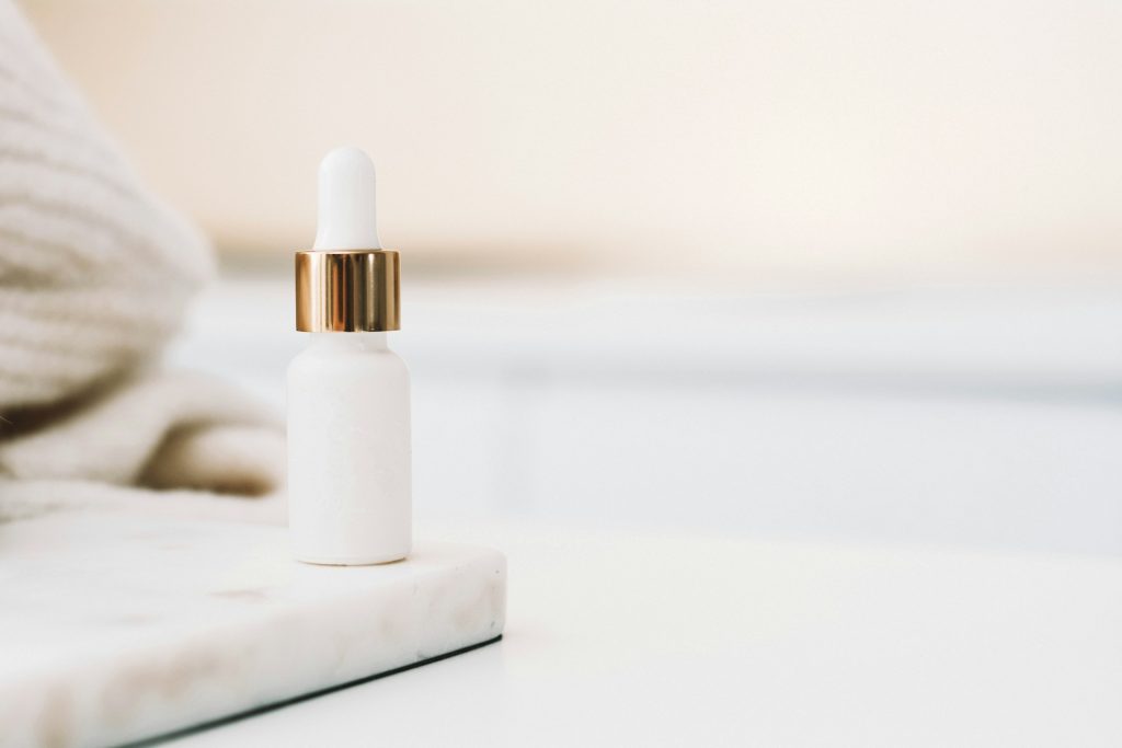 An unlabeled white dropper bottle with a gold lid sits on a white marble surface on a white table.