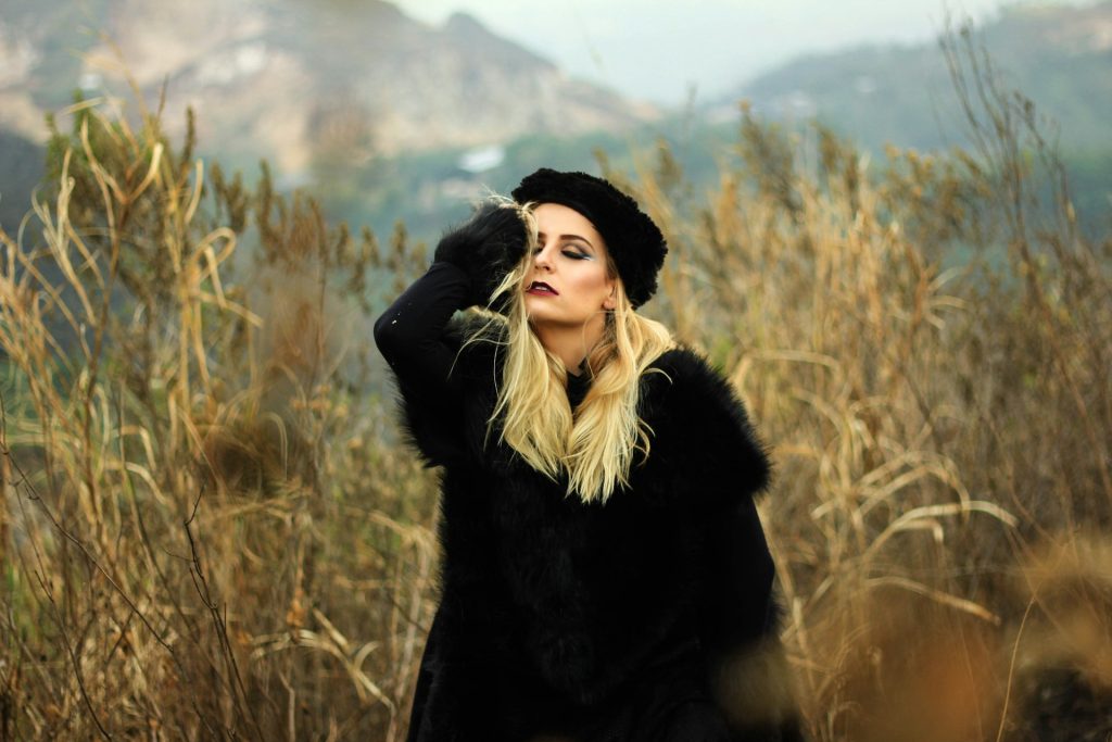 A woman in the mob wife aesthetic stands in a tall grassy field with one hand to her head and her eyes closed. She wears a black fur coat and black fur hat.