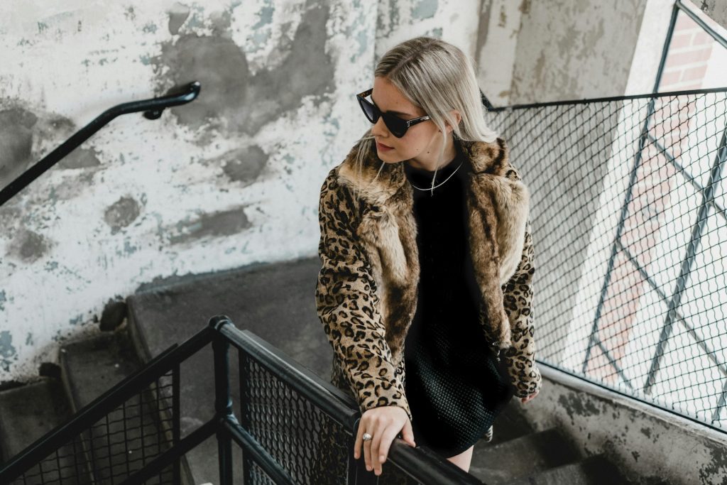 A woman climbs an industrial staircase looking over her shoulder. She wears chunky black sunglasses, a leopard print fur coat, a black dress, a silver ring and a silver necklace.