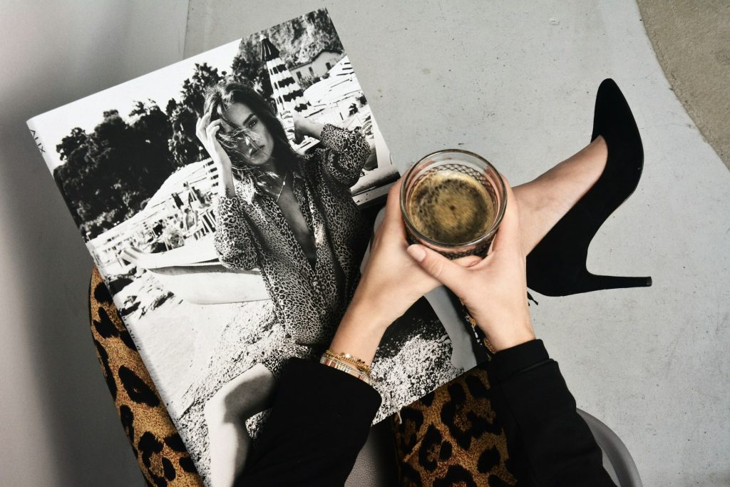 A woman sits with her legs crossed and hands holding a cup of black soda or coffee. She wears a black long-sleeved shirt, gold bracelets and leopard print pants. A black and white magazine sits on her lap with a model wearing leopard print pajamas by a boat. The woman holding the magazine also wears black stilettos.
