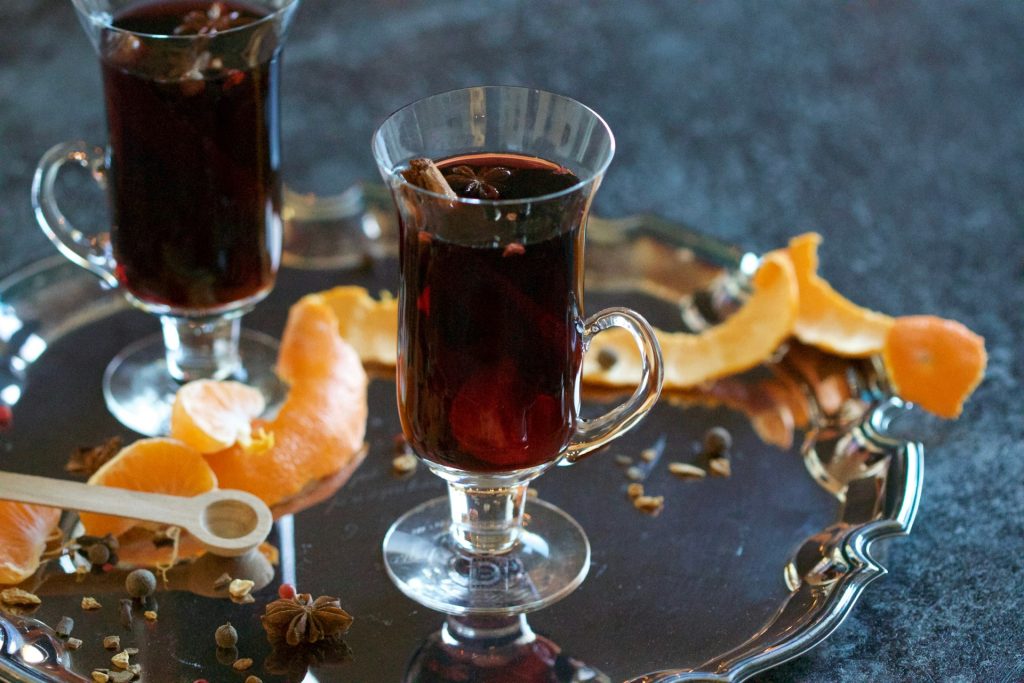 Two glasses containing mulled wine sit on a silver platter among orange rinds and scattered herbs