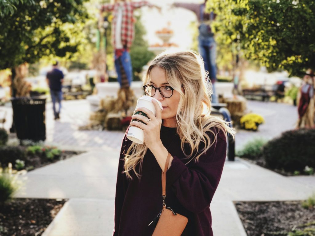 A woman with recession blonde hair sips from a Starbucks cup in the middle of a fall festival. She wears a burgundy pullover sweater and has a brown leather clutch hanging from a strap around her wrist. She wears black round glasses and a gold ring.