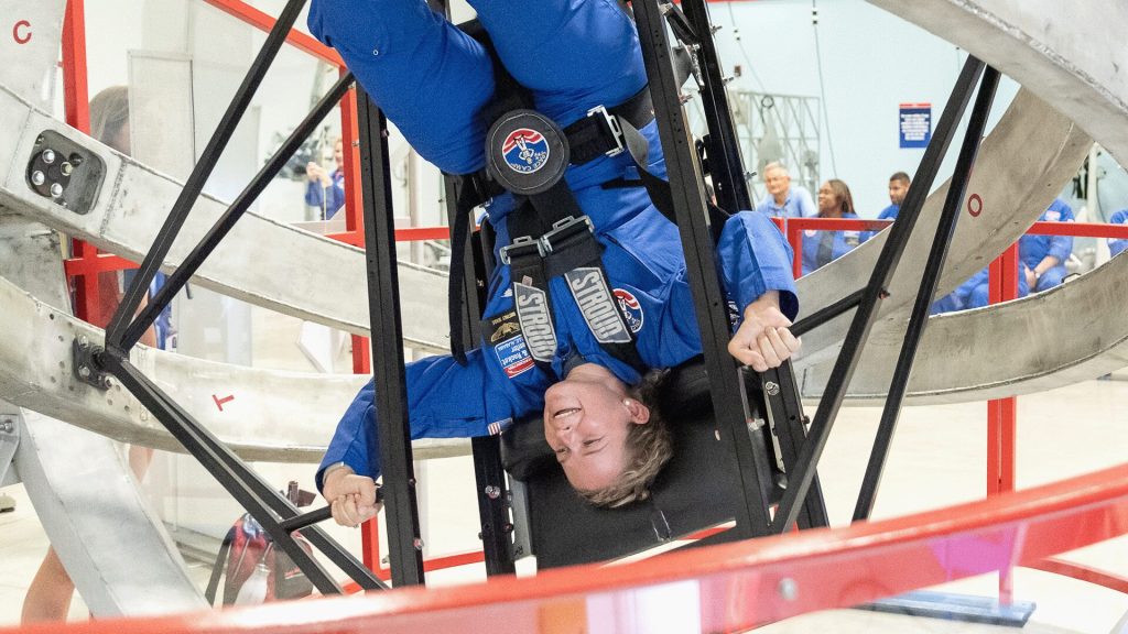 A woman hangs upside down in rotating equipment used to train astronauts. She wears a blue astronaut suit like the people watching behind her.