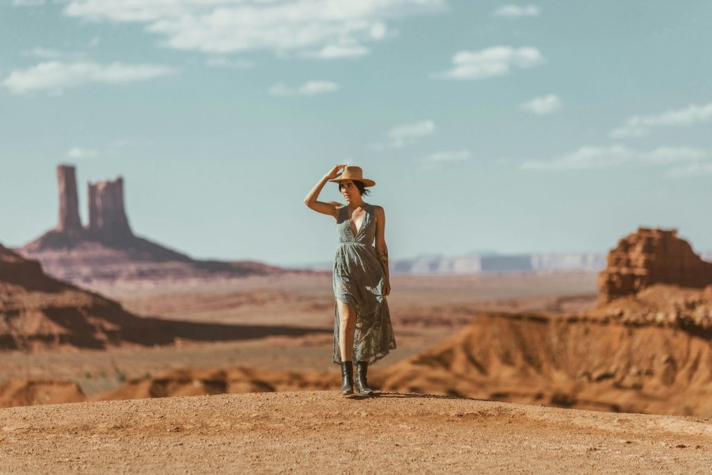 A woman stands in a light green midi sundress with a deep v-neck, black boots and a straw hat. She's in the middle of a desert under a cloudy blue sky.