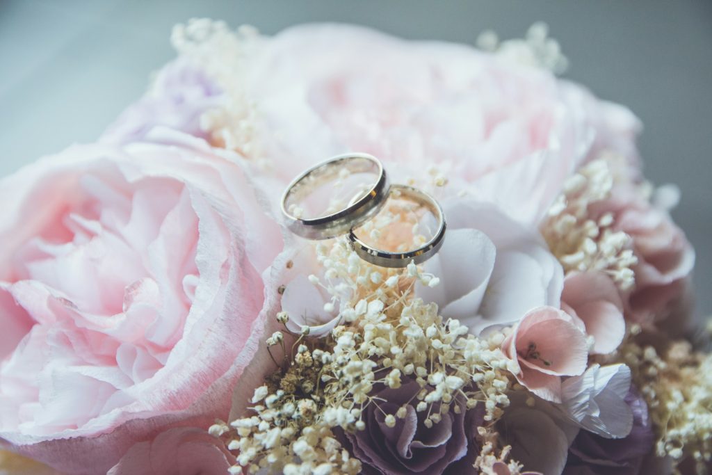 Two silver wedding bands sit on a bouquet of pink peonies and white baby's breath flowers.