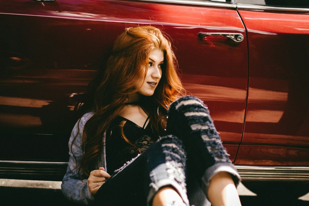 A redheaded woman with long hair wearing the whimsigoth aesthetic clothing sits against a red car door.