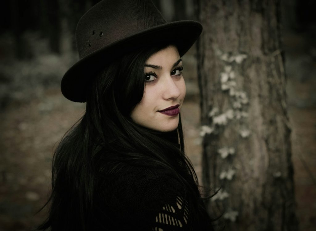 A woman in whimsigoth clothing with a black hat looks over her shoulder while in the woods.