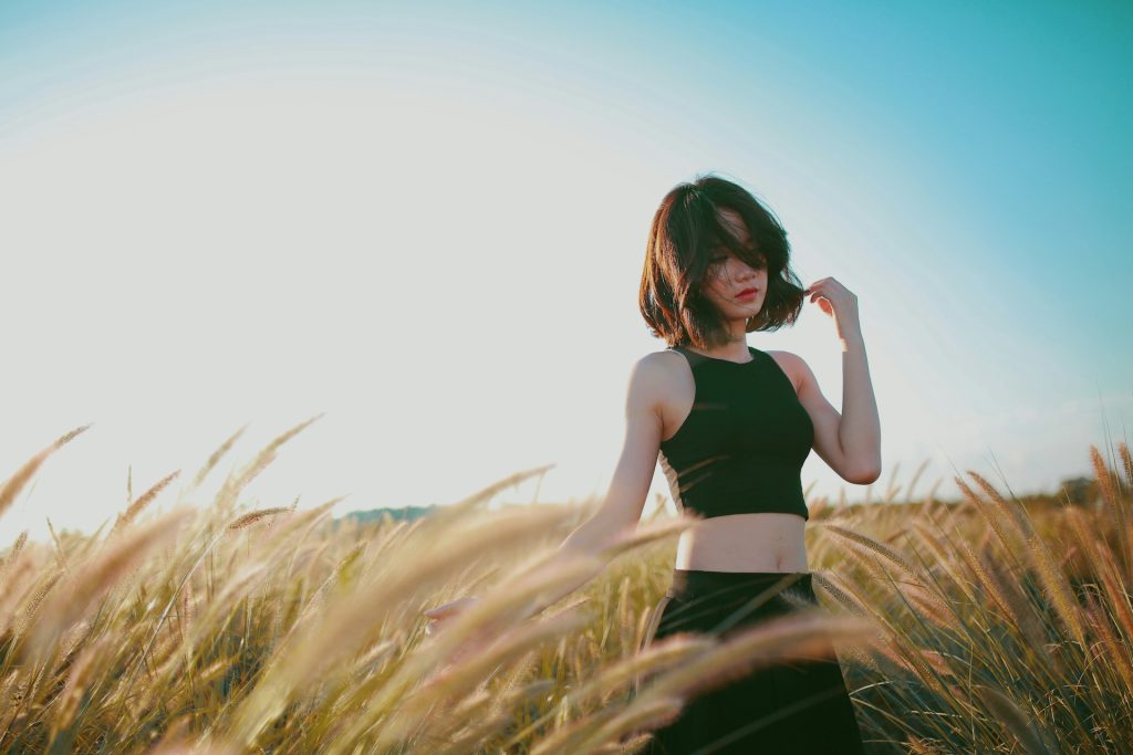 A woman stands in a black crop top tank top and black pants in a wheat field under a blue sky.