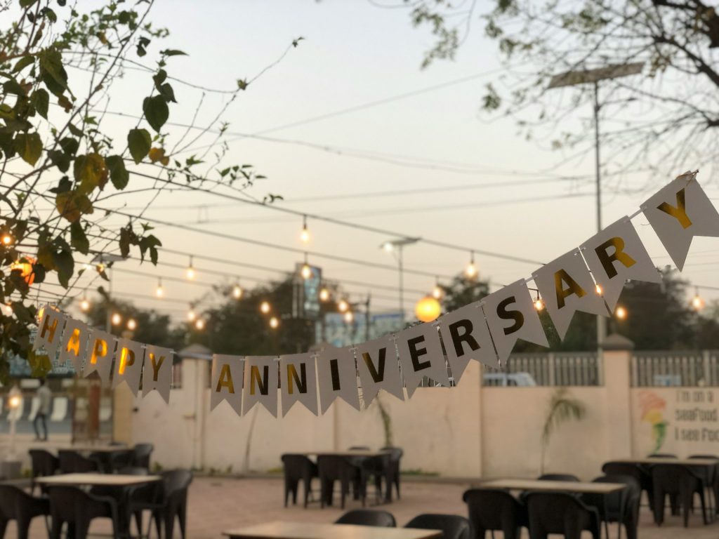 A white and gold Happy Anniversary banner hangs over outdoor seating. The tables and chairs are surrounded by a white wall, trees, a blue sky and glowing Edison bulbs over the dining area.