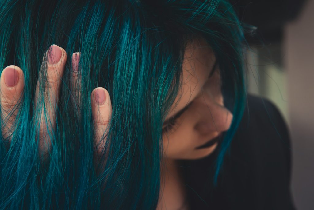 A woman runs her fingers through her short teal hair. She wears black lipstick and leans her head toward the camera.