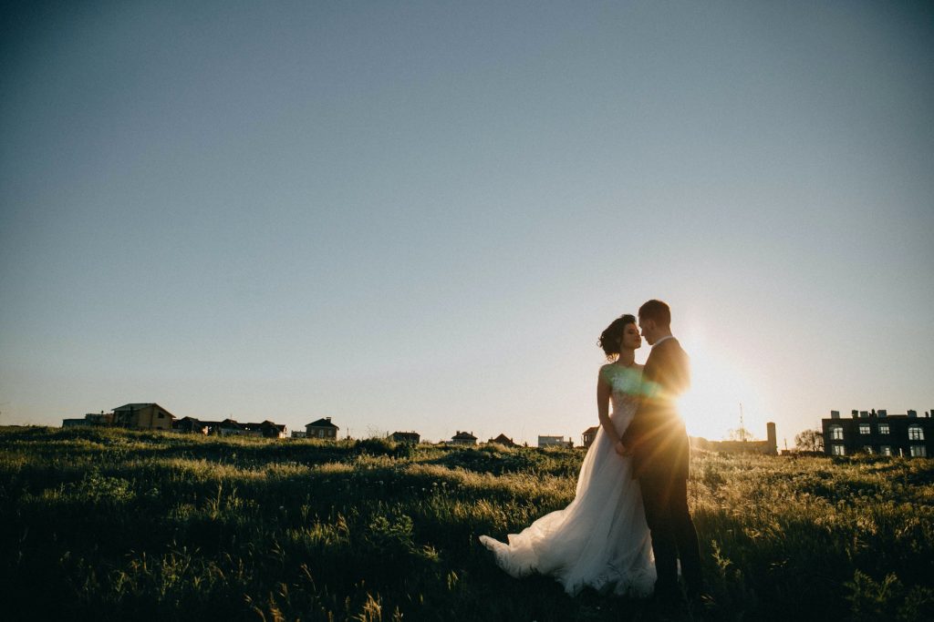 A bride and groom stands in a field at sunset.
