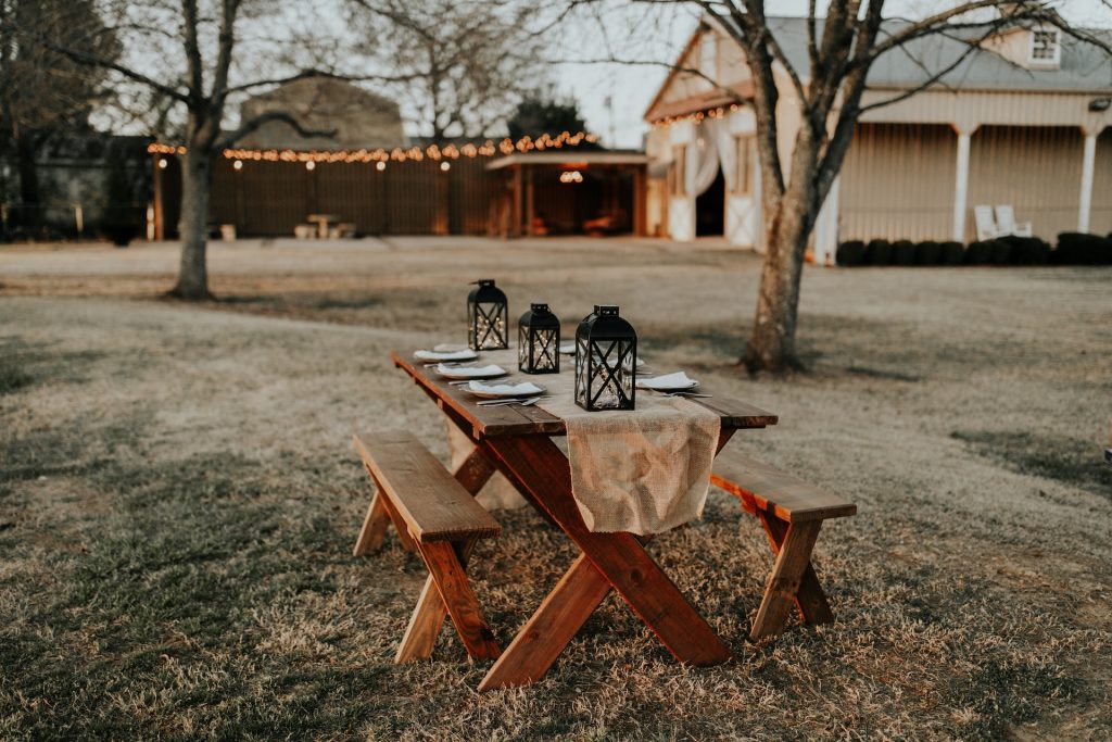 A picnic table outside of a barn is set with a table cloth and lanterns.