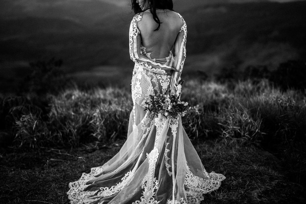 A woman wears a sheer wedding dress and holds a bouquet behind her back.