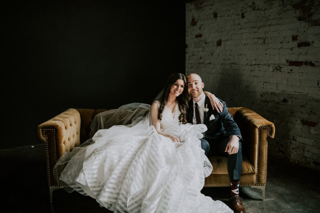 A bride wears a striped, ruffled gown, sitting next to a groom in a suit.