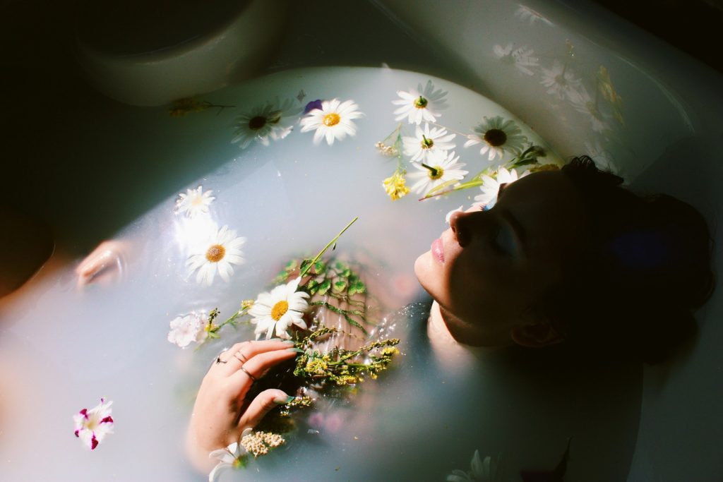 A woman relaxes in a white bath full of daisies, likely because she's doing a spiritual bath cleanse.