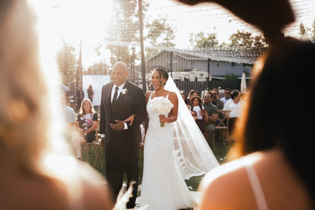 A father walks a bride down the aisle at an outdoor wedding ceremony.