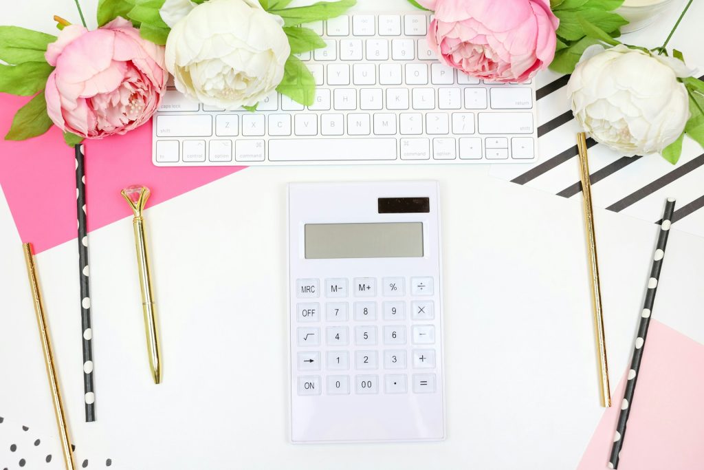 A white calculator sits on a white desk surrounded by white and pink peonies, pens and pencils. A white Macbook keyboard sits above the calculator. Both can help people find their life path number.