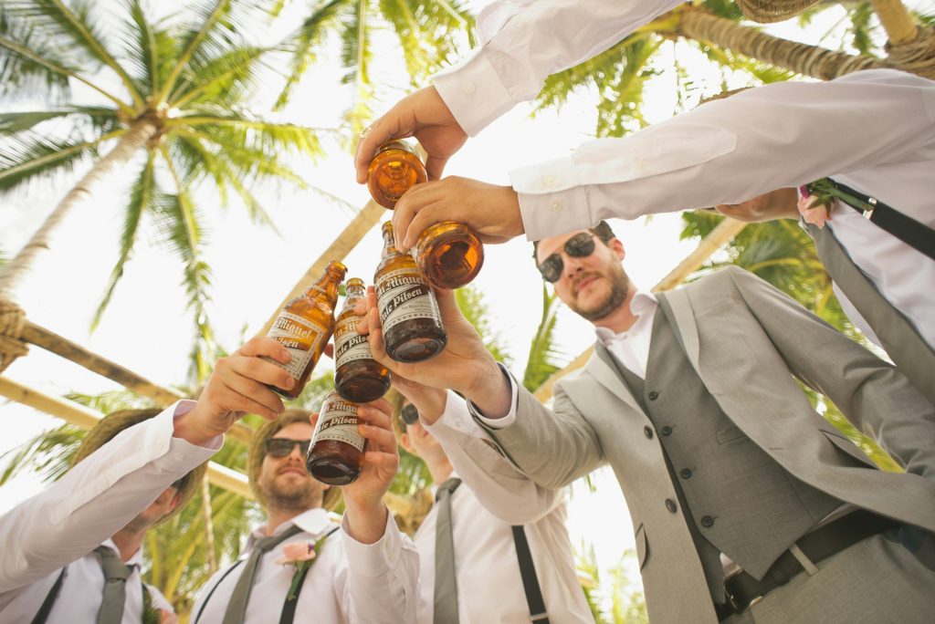 A group of men in wedding party attire clink beer bottles together under palm trees.