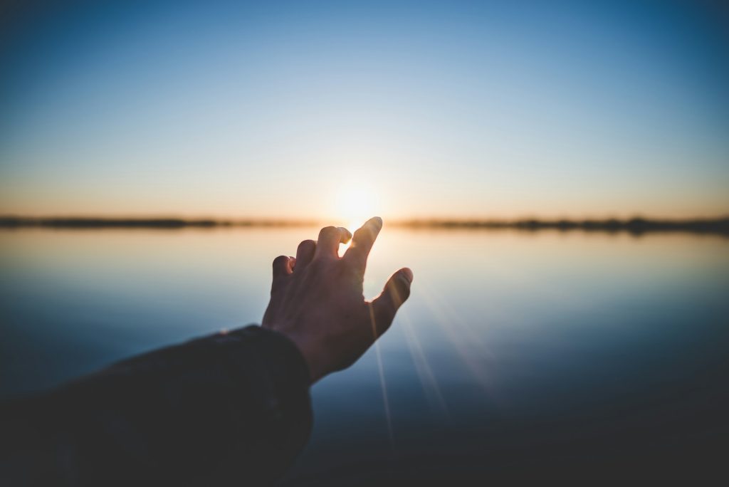 A person in a long sleeved black shirt reaches out toward the sun rise. An expansive lake sits in front of them under a clear blue sky.