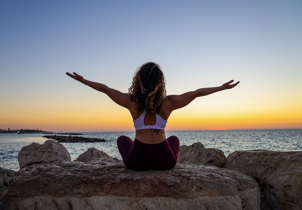 A woman sits with her arms raised on a rock overlooking the ocean.