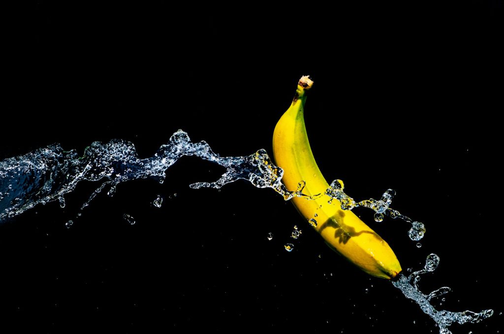 A bright yellow and light green banana sits against a black background. A splash of water cascades over the banana.