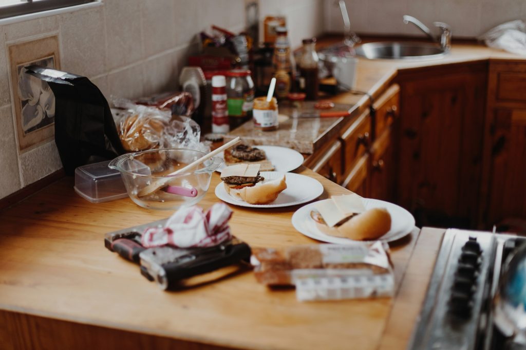 A light brown wooden kitchen counter is covered in ingredients while someone preps cheeseburgers.